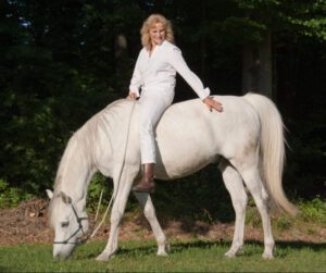 Horsemanship Impressum: Ingrid Merkle sitzt auf einem Araber-Pferd Schimmel und streichelt ihn. Das Pferd frisst Gras.