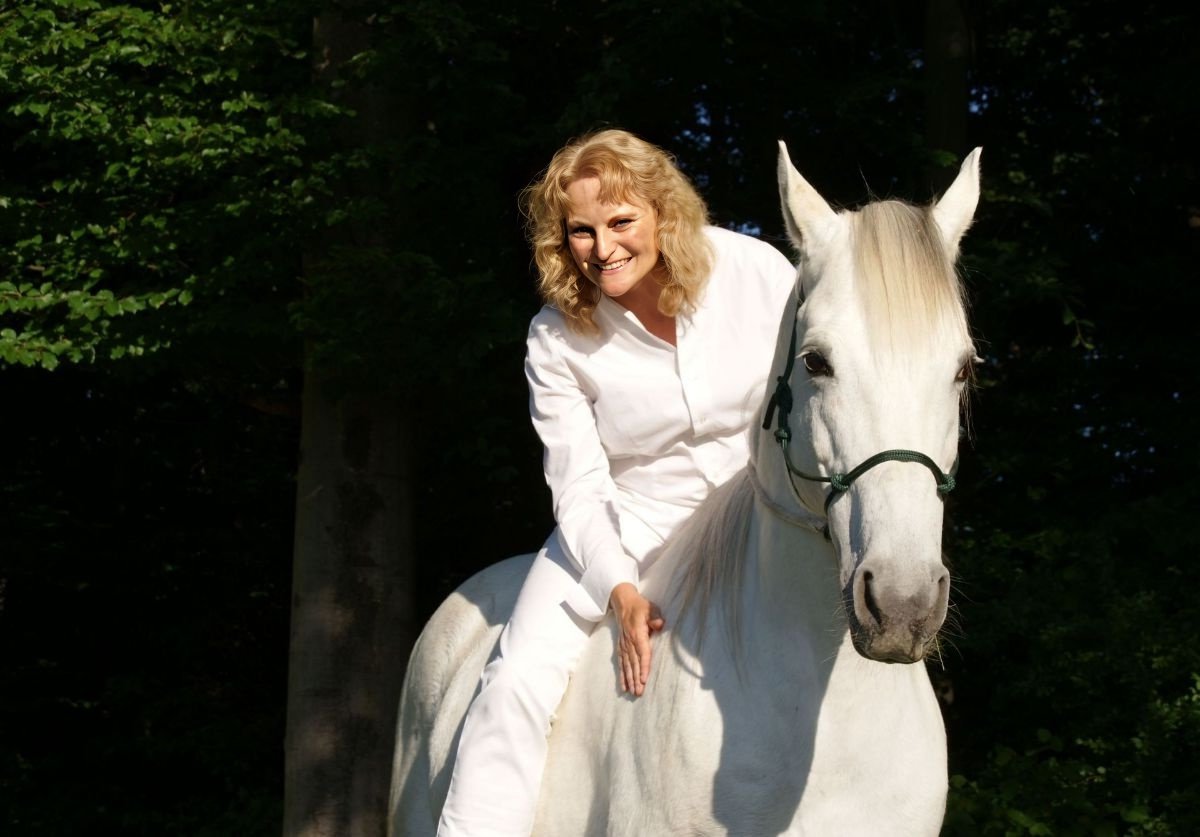 Horsemanship Academy Gründerin Ingrid Merkle reitet auf ihrem Schimmel Araber-Pferd im Wald und streichelt ihn, sie lächelt.